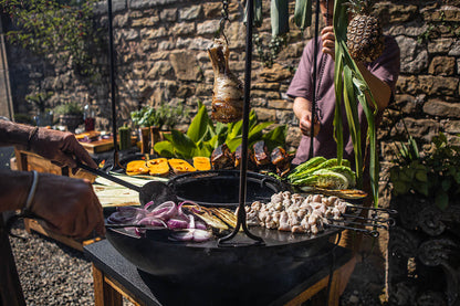 Outdoor plancha kadai grill cooking vegetables, skewers and hanging meat over open fire