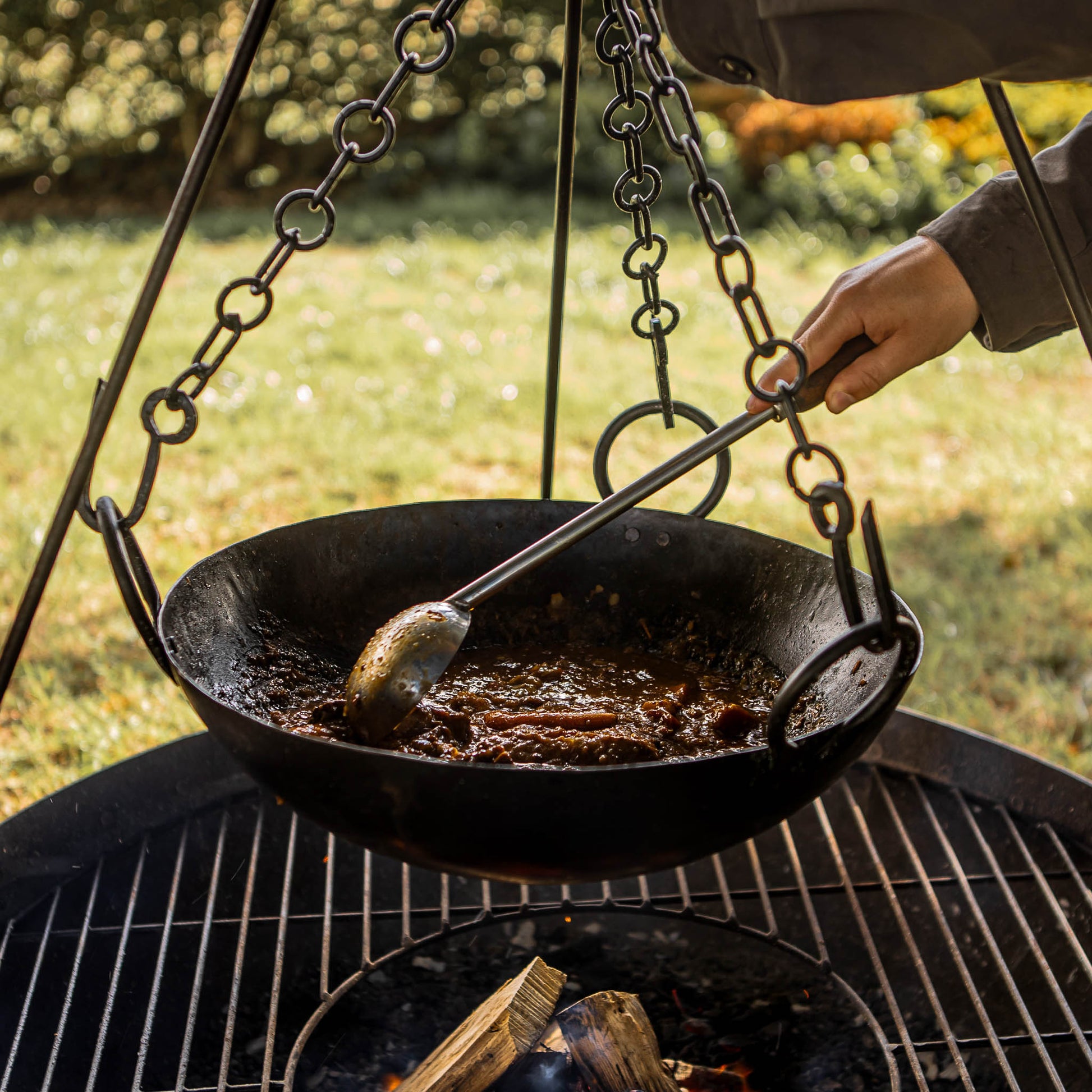 Stainless steel barbecue ladle used for serving and stirring fire-cooked dishes in a suspended outdoor cooking bowl