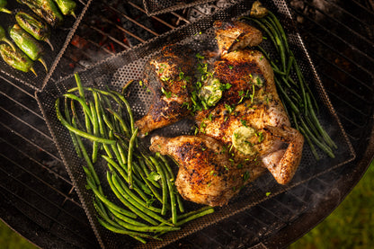 Spatchcocked chicken and grilled green beans cooking on a perforated BBQ grill tray over open coals, with charred peppers visible nearby and melted herb butter on the chicken.