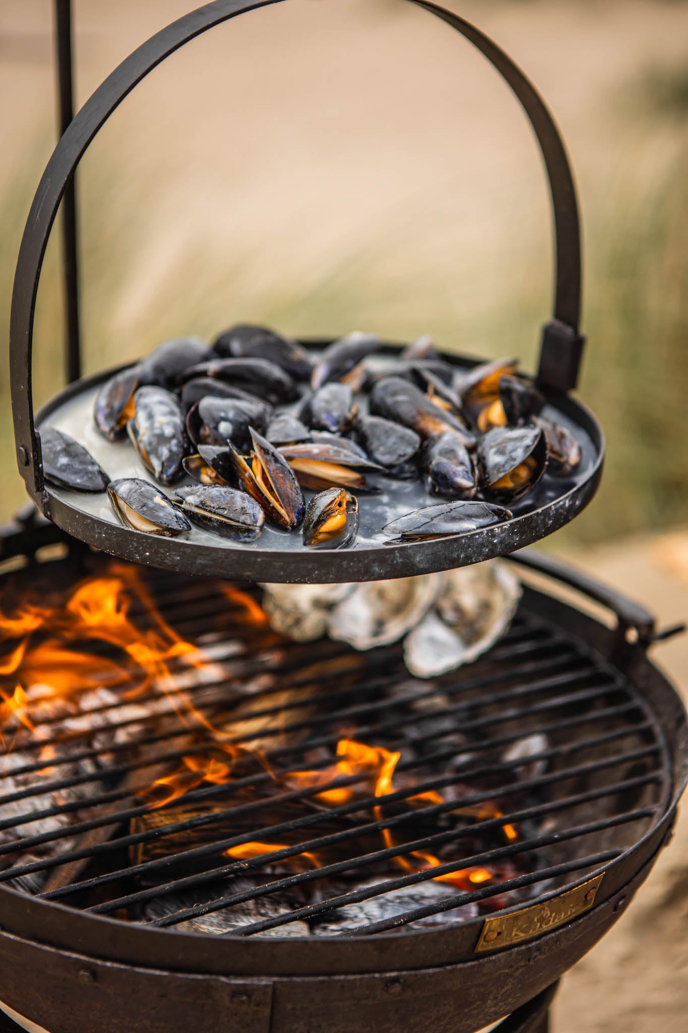 Portable BBQ skillet setup with mussels on a hanging rack above a Kadai grill over open flames
