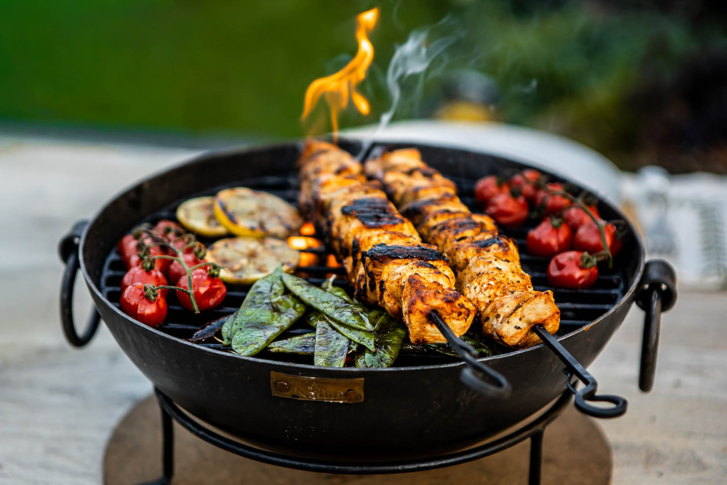 Outdoor BBQ table with Kadai fire bowl cooking skewers, tomatoes and greens with flames rising