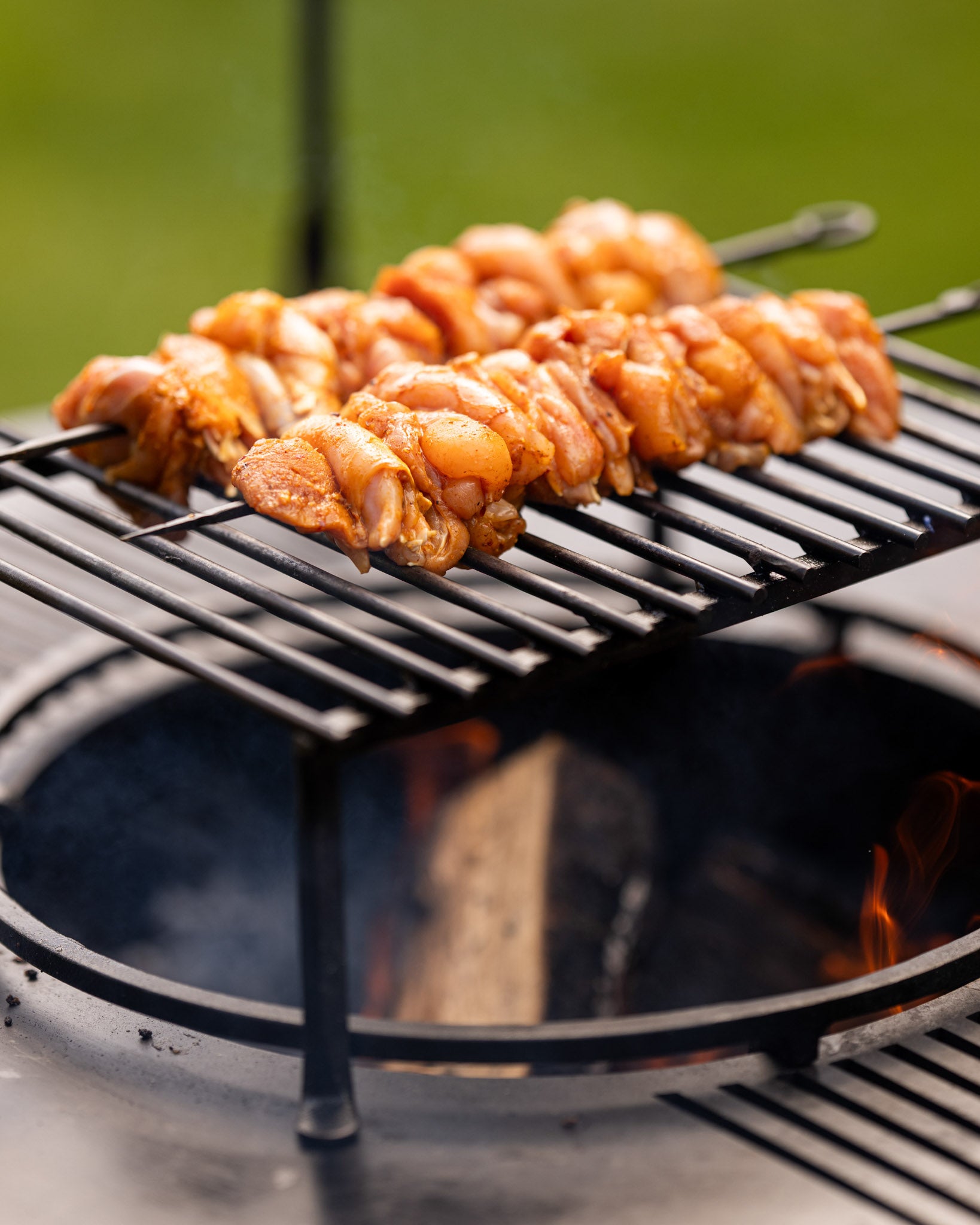 Marinated chicken BBQ skewers grilling over an open flame on a metal rack above a fire pit.