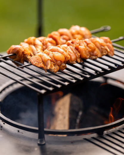 Marinated chicken BBQ skewers grilling over an open flame on a metal rack above a fire pit.