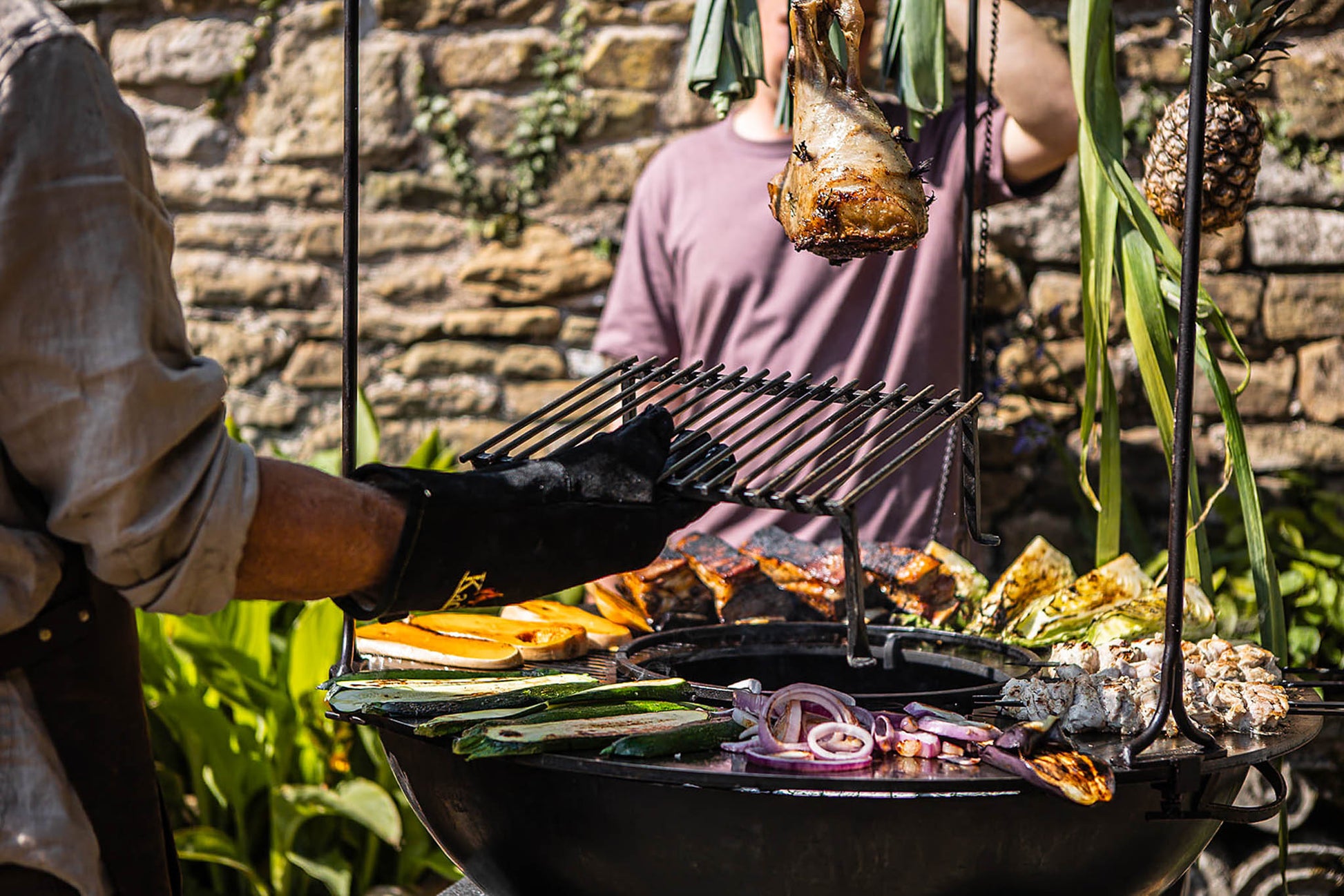 Outdoor table grill with vegetables cooking and meat hanging above the fire