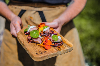 Handcrafted teak chopping board used to serve grilled bacon bites garnished with edible flowers and fresh green leaves outdoors
