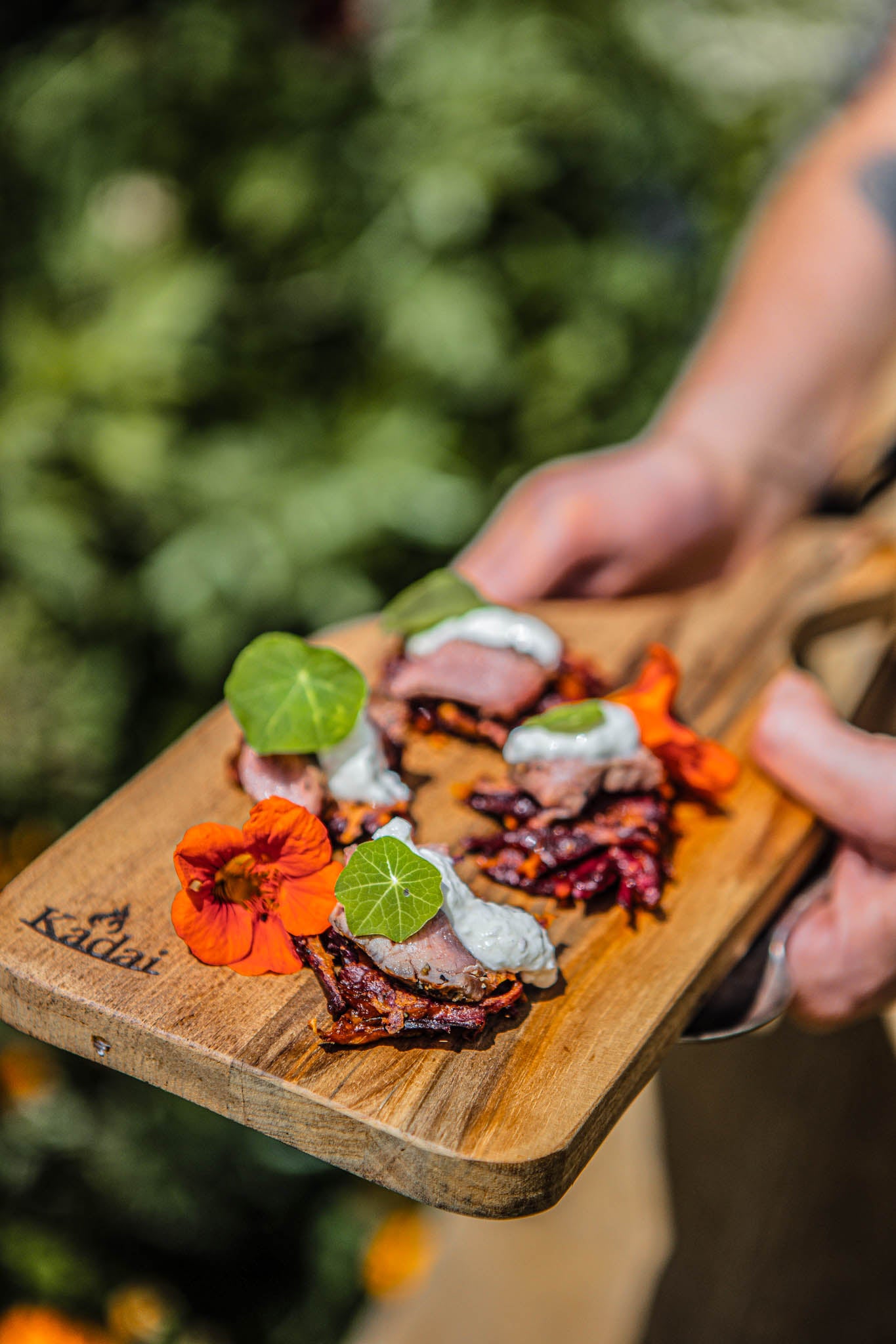 Close-up of a handcrafted teak serving board presenting elegant barbecue canapés with colourful floral garnish and creamy topping