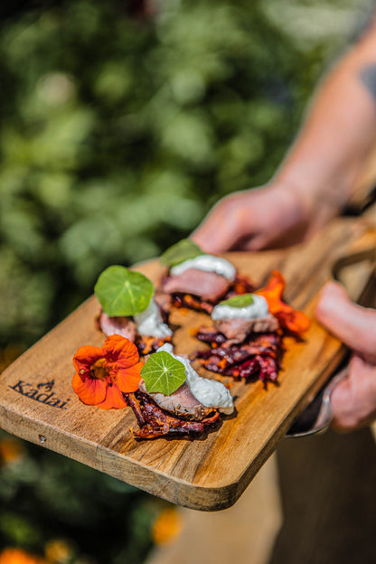 Close-up of a handcrafted teak serving board presenting elegant barbecue canapés with colourful floral garnish and creamy topping