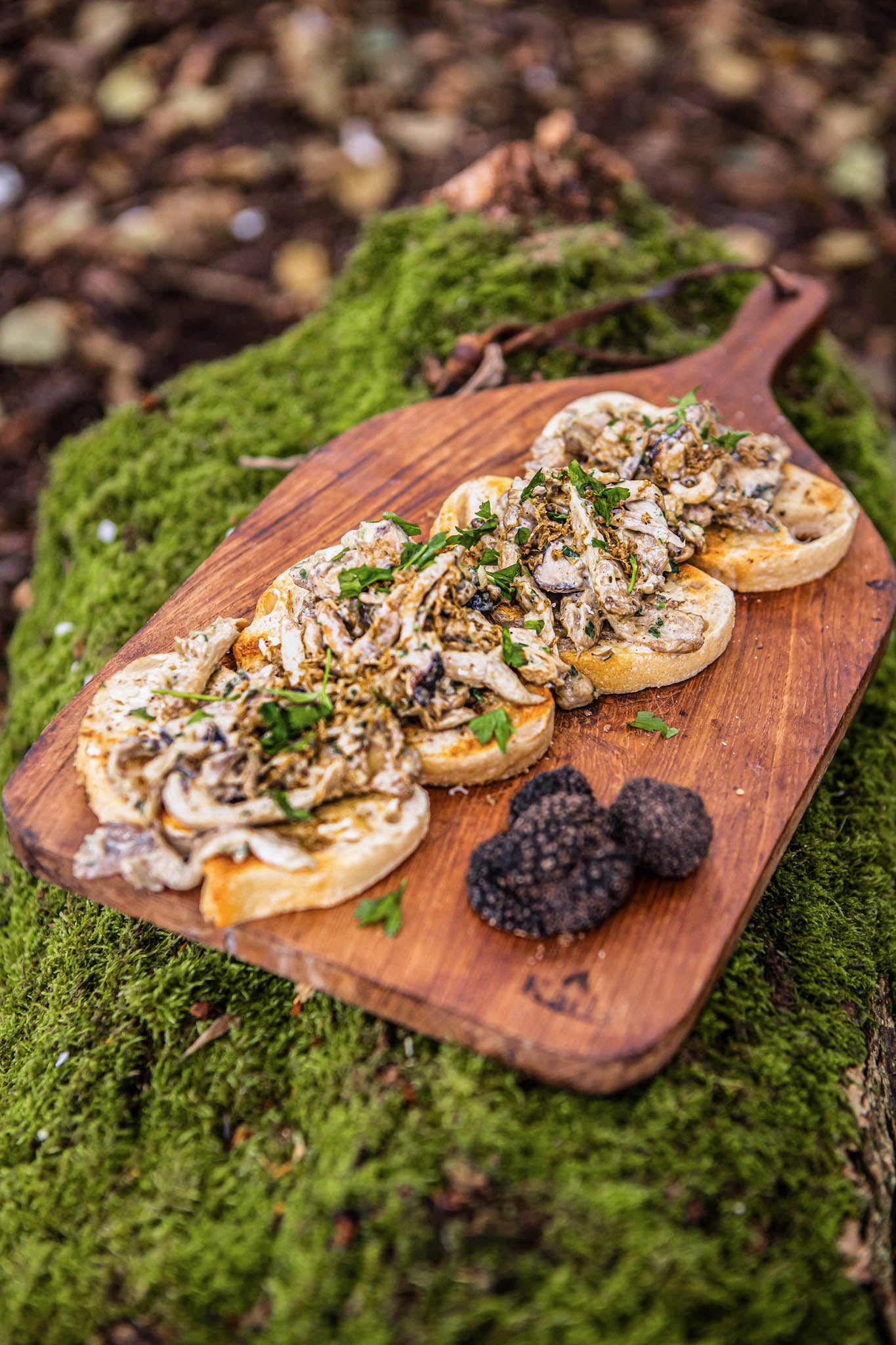 Handcrafted teak chopping board displaying mushroom crostini and truffles on a moss-covered woodland surface