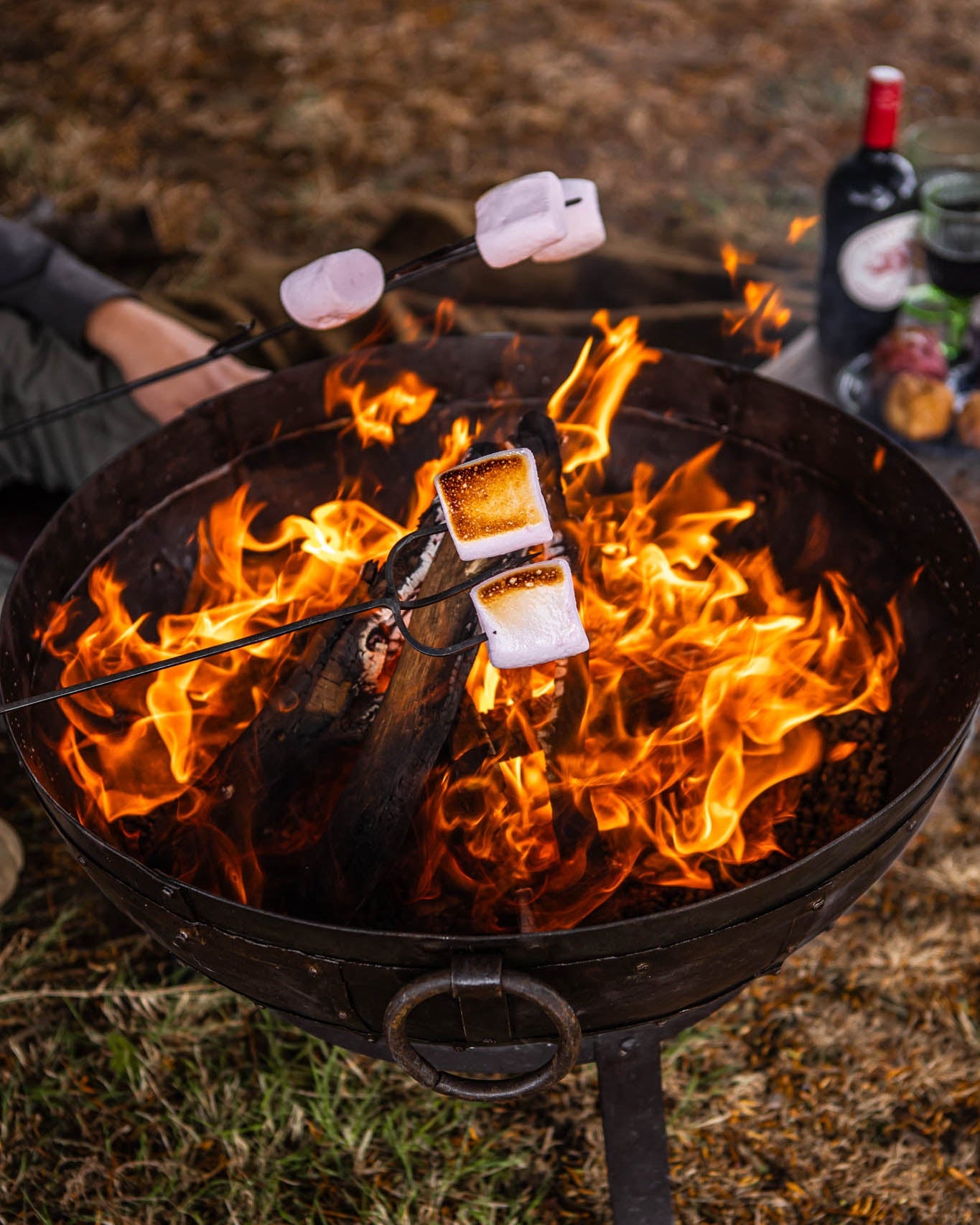 Marshmallows roasting on long metal toasting forks above a blazing outdoor fire pit with glowing flames and logs.