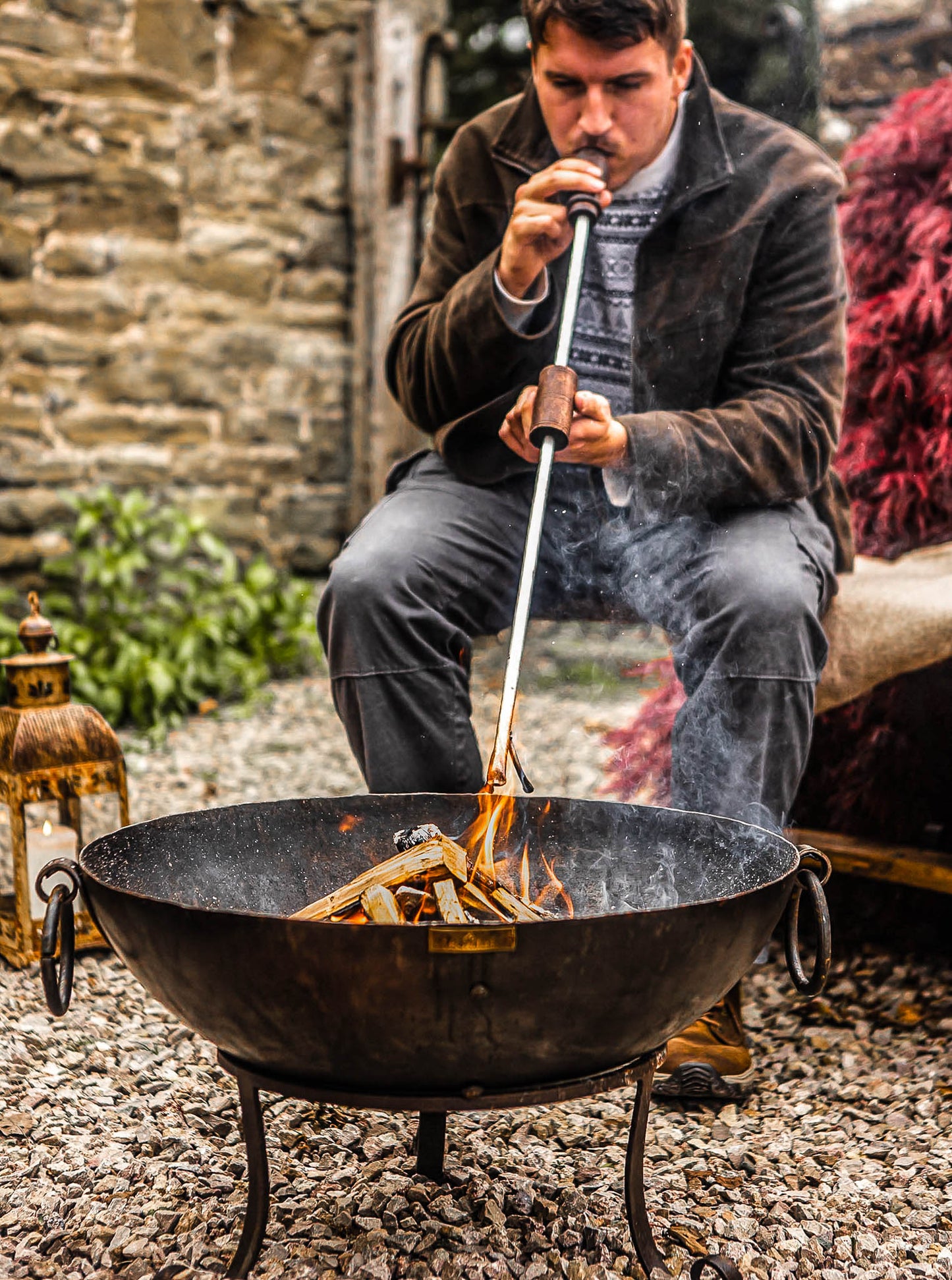 Man using a long metal blow poker with wooden handles to revive flames in a Kadai fire bowl filled with kindling, set on a gravel patio beside rustic outdoor seating.