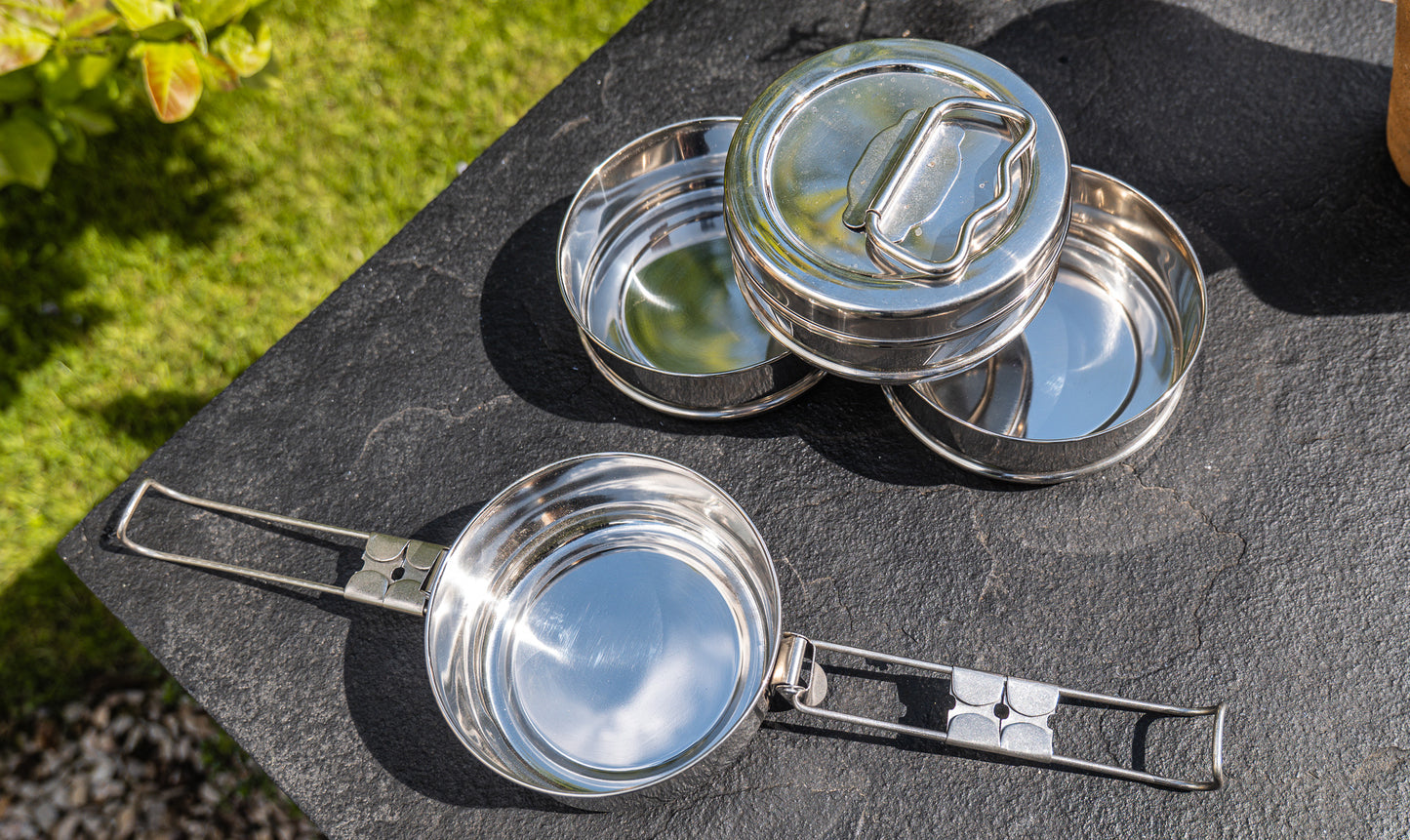 Set of metal tiffin tin containers on a stone surface with greenery in the background