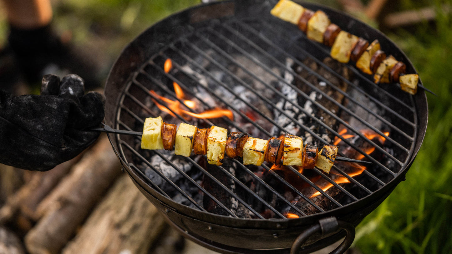 Skewer of pineapple and sausage cooking over open flames on a portable BBQ skillet grill