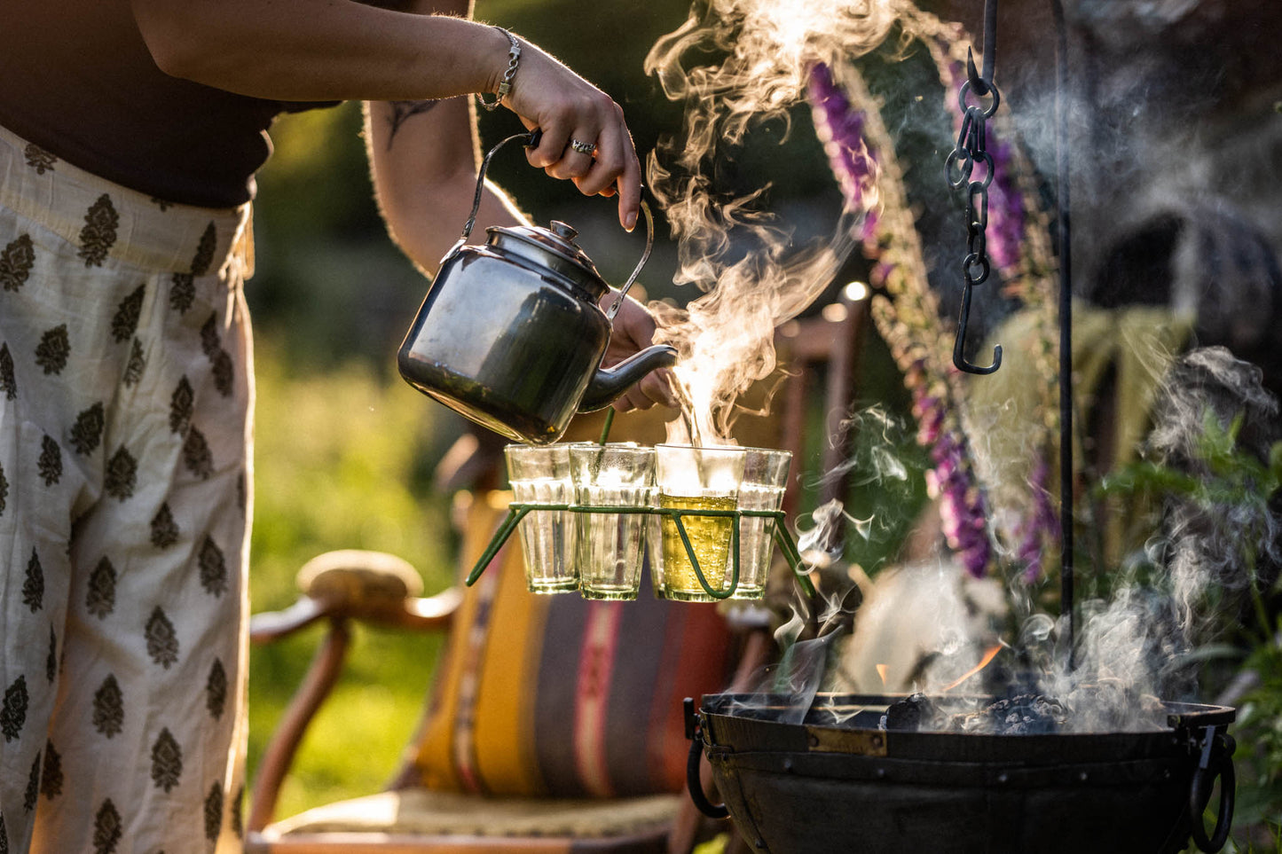Camping BBQ kettle pouring hot tea into glasses beside a fire pit, with steam rising in an outdoor garden setting
