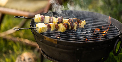 Portable BBQ with Kadai grill cooking skewers of pineapple and sausage over charcoal with flames and smoke