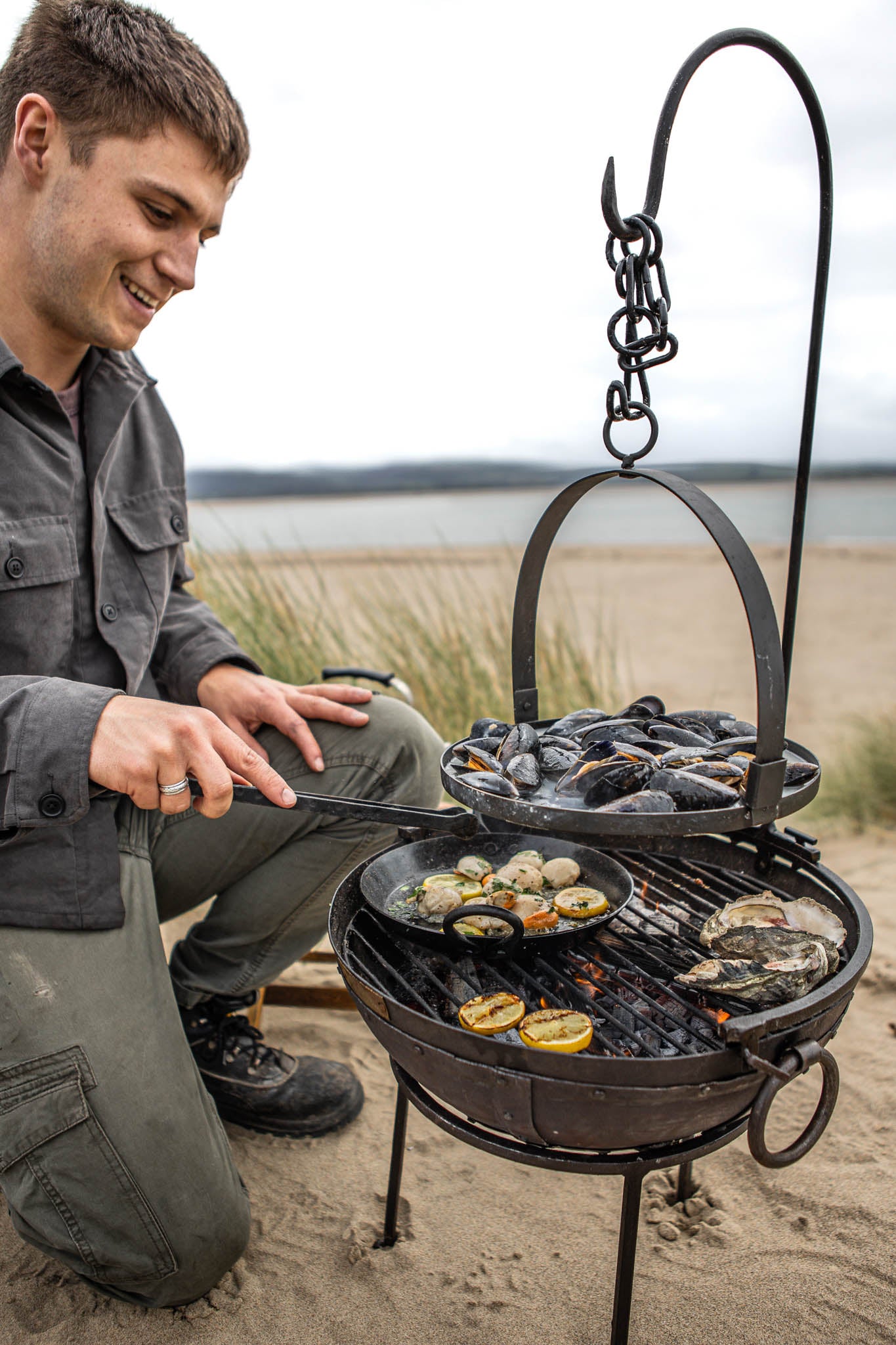 Portable BBQ fire pit with Kadai grill cooking seafood on a beach, including mussels, scallops and oysters