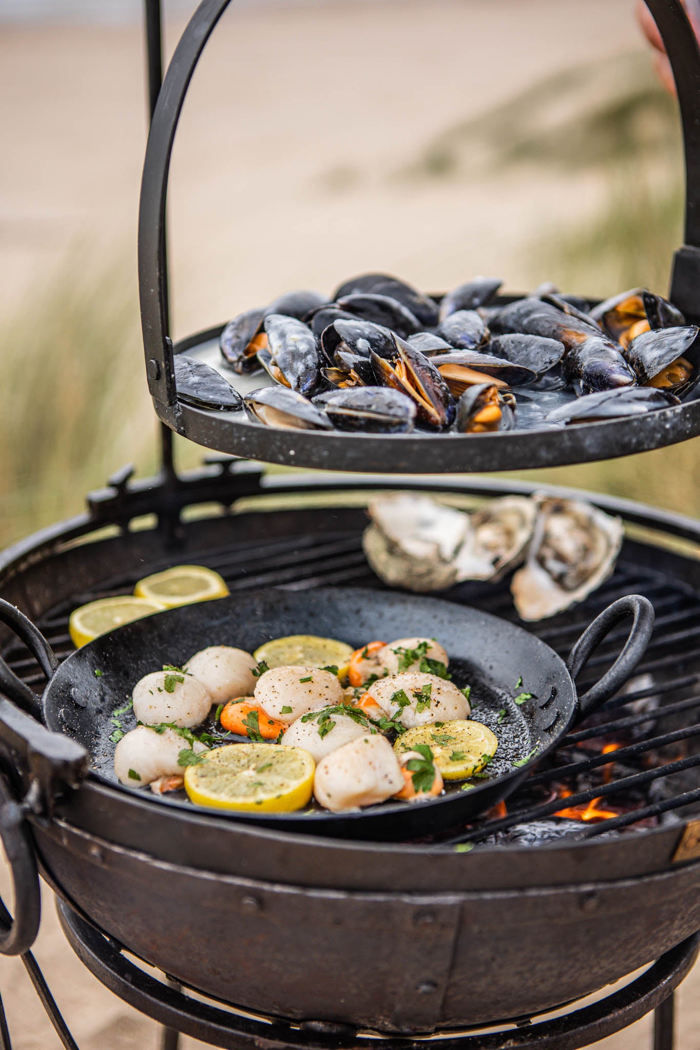 Portable BBQ skillet cooking scallops in a pan on a Kadai grill, with mussels on a raised rack above the fire