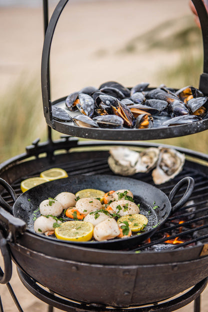 Portable BBQ skillet cooking scallops in a pan on a Kadai grill, with mussels on a raised rack above the fire