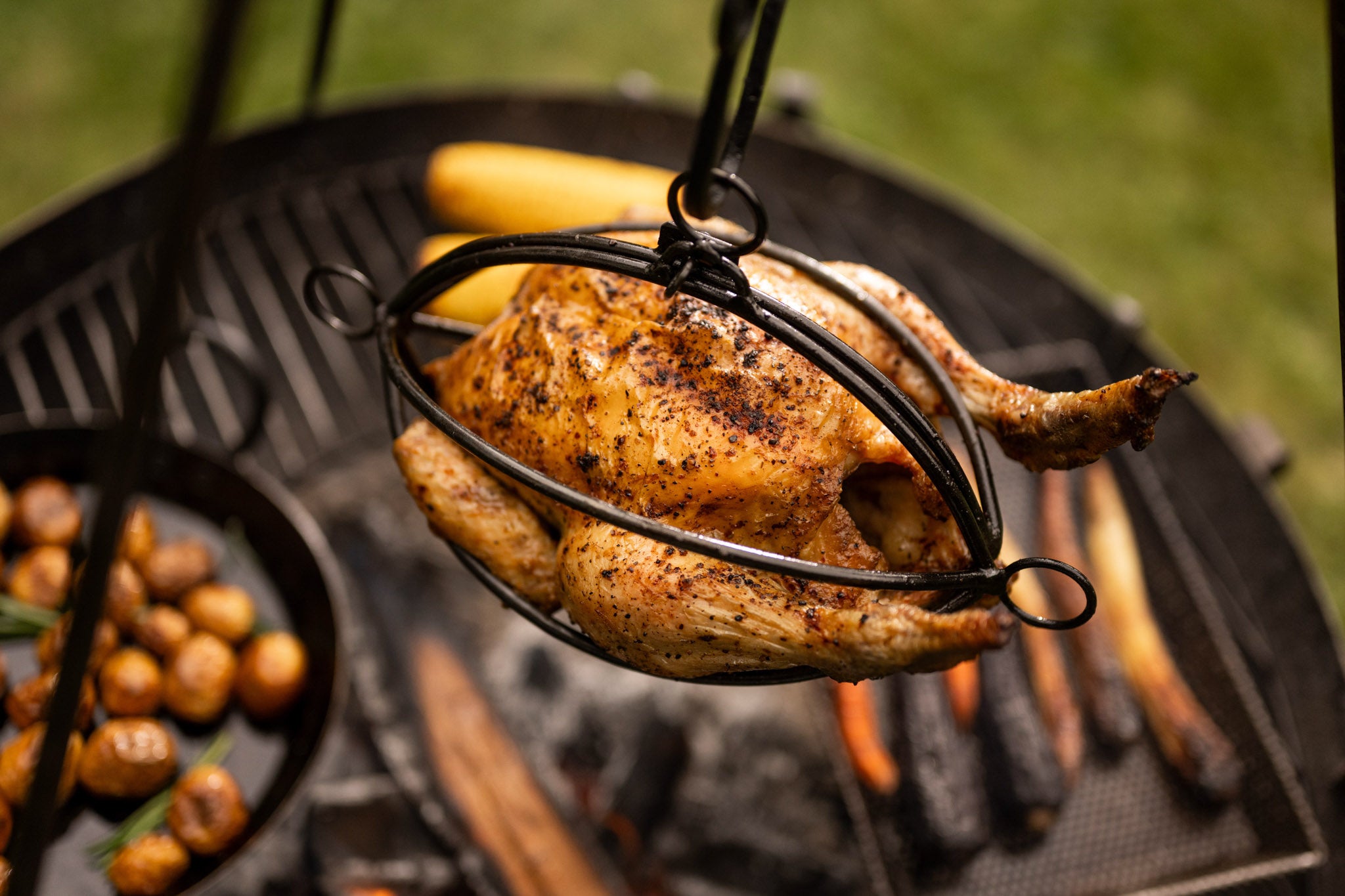 Whole roasted chicken hanging in a BBQ tripod roaster basket above glowing firewood and outdoor grill coals.