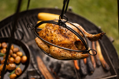Whole roasted chicken hanging in a BBQ tripod roaster basket above glowing firewood and outdoor grill coals.
