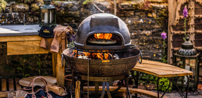 Wood fired oven on a Kadai fire pit with visible flames cooking pizza in an outdoor garden setting