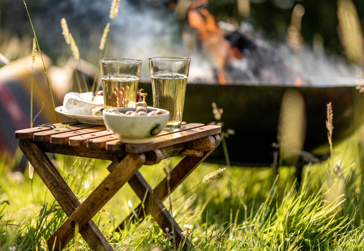 Foldable camping stool used as a small outdoor table with drinks and snacks beside a fire pit